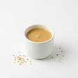 A white ceramic cup filled with smooth peanut butter, photographed on a clean white background with a few sesame seeds scattered nearby.