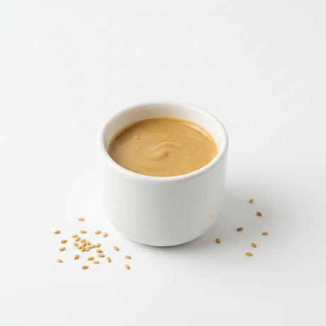 A white ceramic cup filled with smooth peanut butter, photographed on a clean white background with a few sesame seeds scattered nearby.